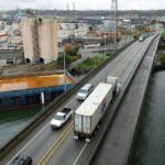 Several cars and a large truck travel on a bridge over a waterway with buildings and a barge in the background.