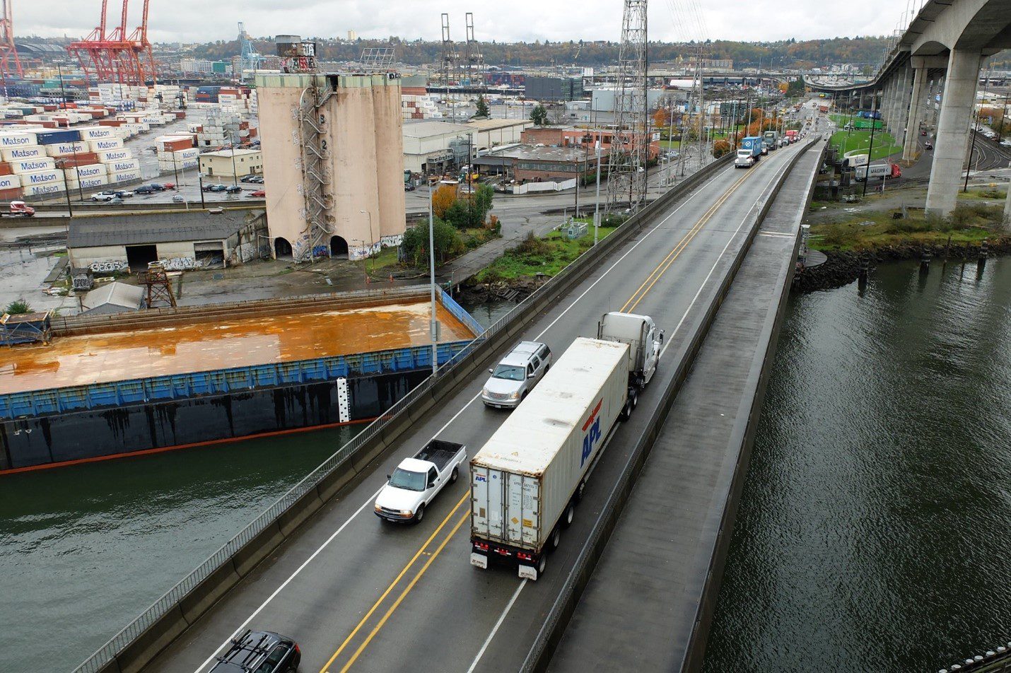 Several cars and a large truck travel on a bridge over a waterway with buildings and a barge in the background.
