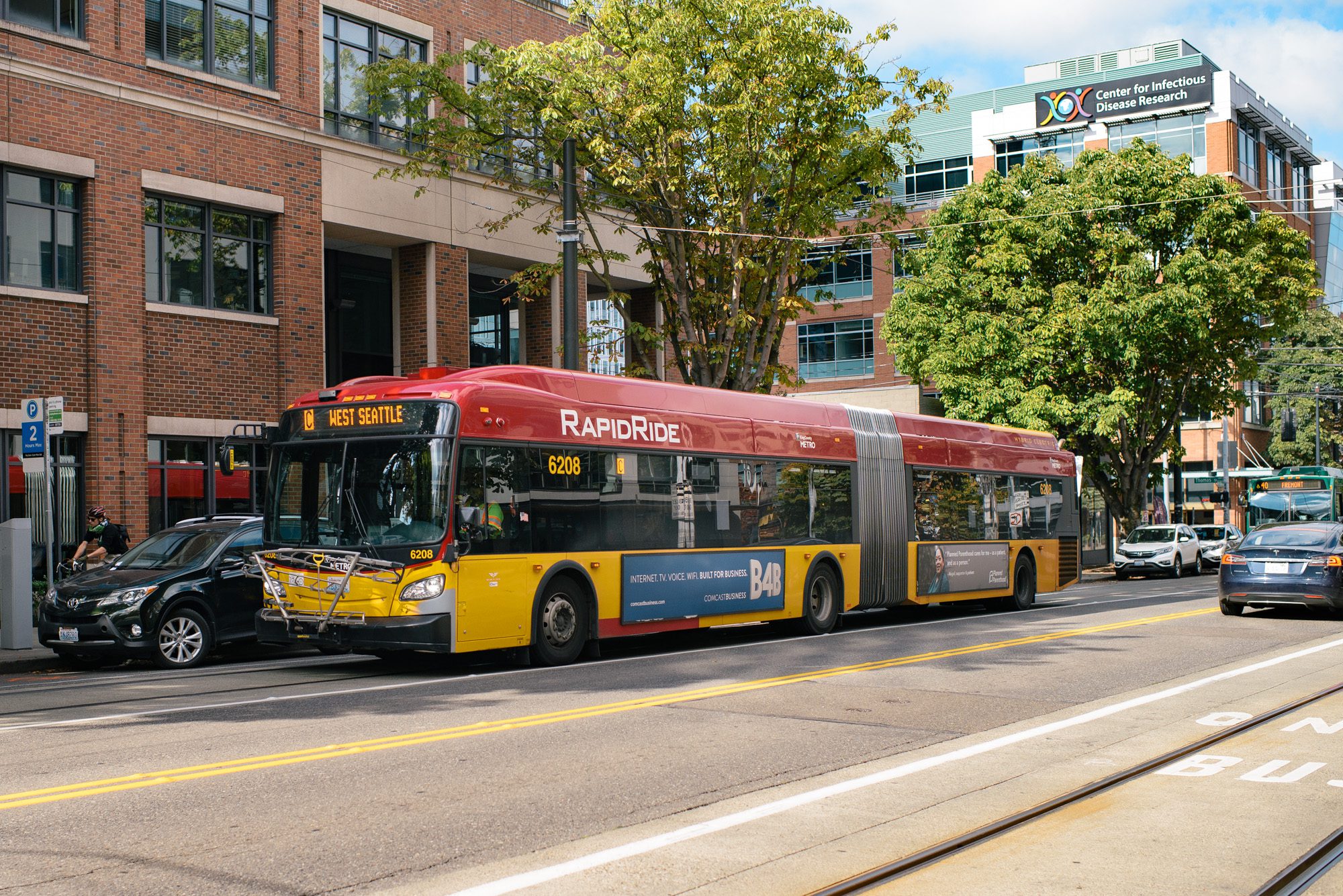 A RapidRide bus travels down a street on a sunny day. Other vehicles, large buildings, and trees are also in the picture.