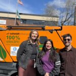 Three people stand together outside in front of a large orange electric sweeper on a sunny day.
