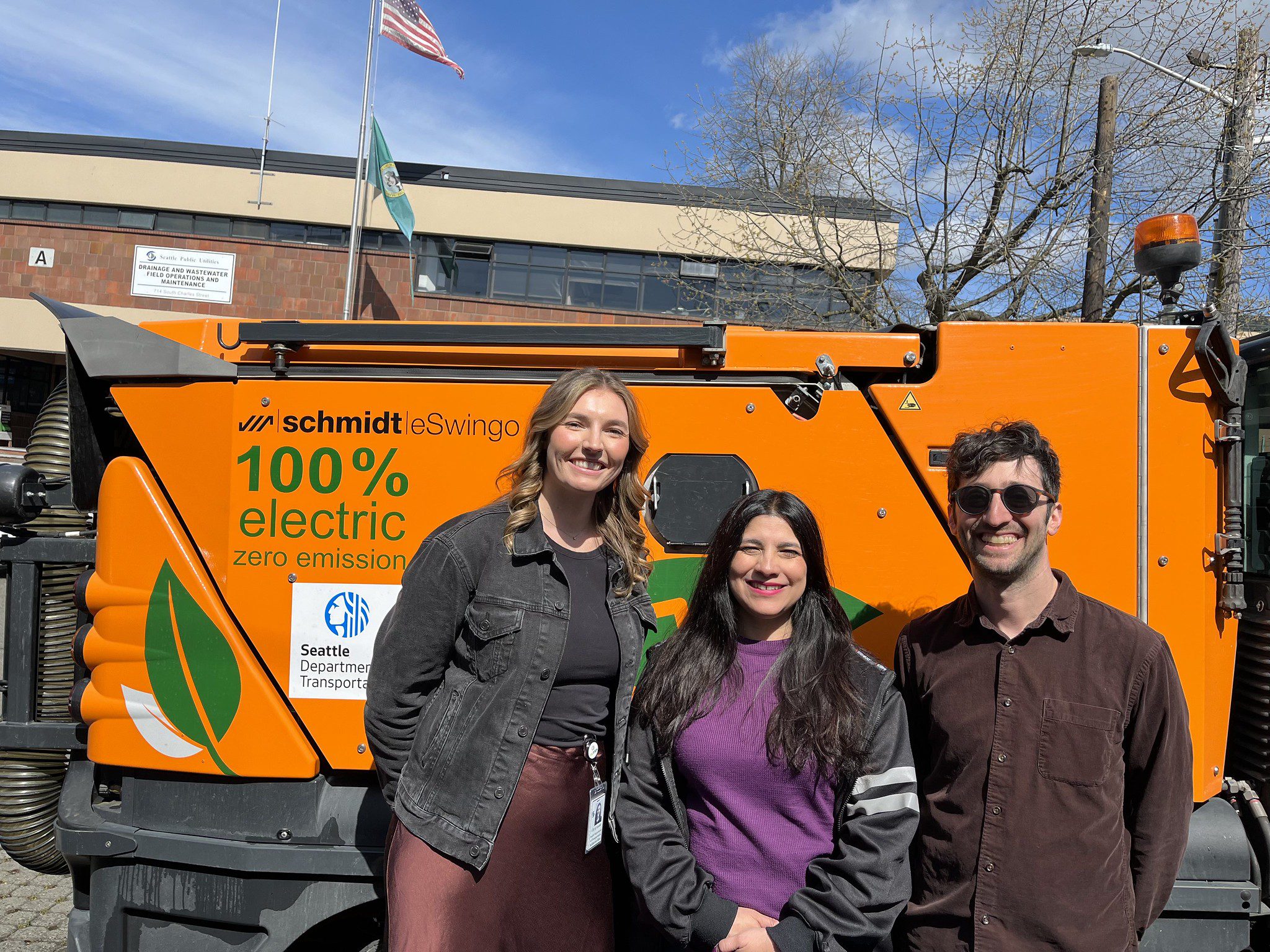 Three people stand together outside in front of a large orange electric sweeper on a sunny day.