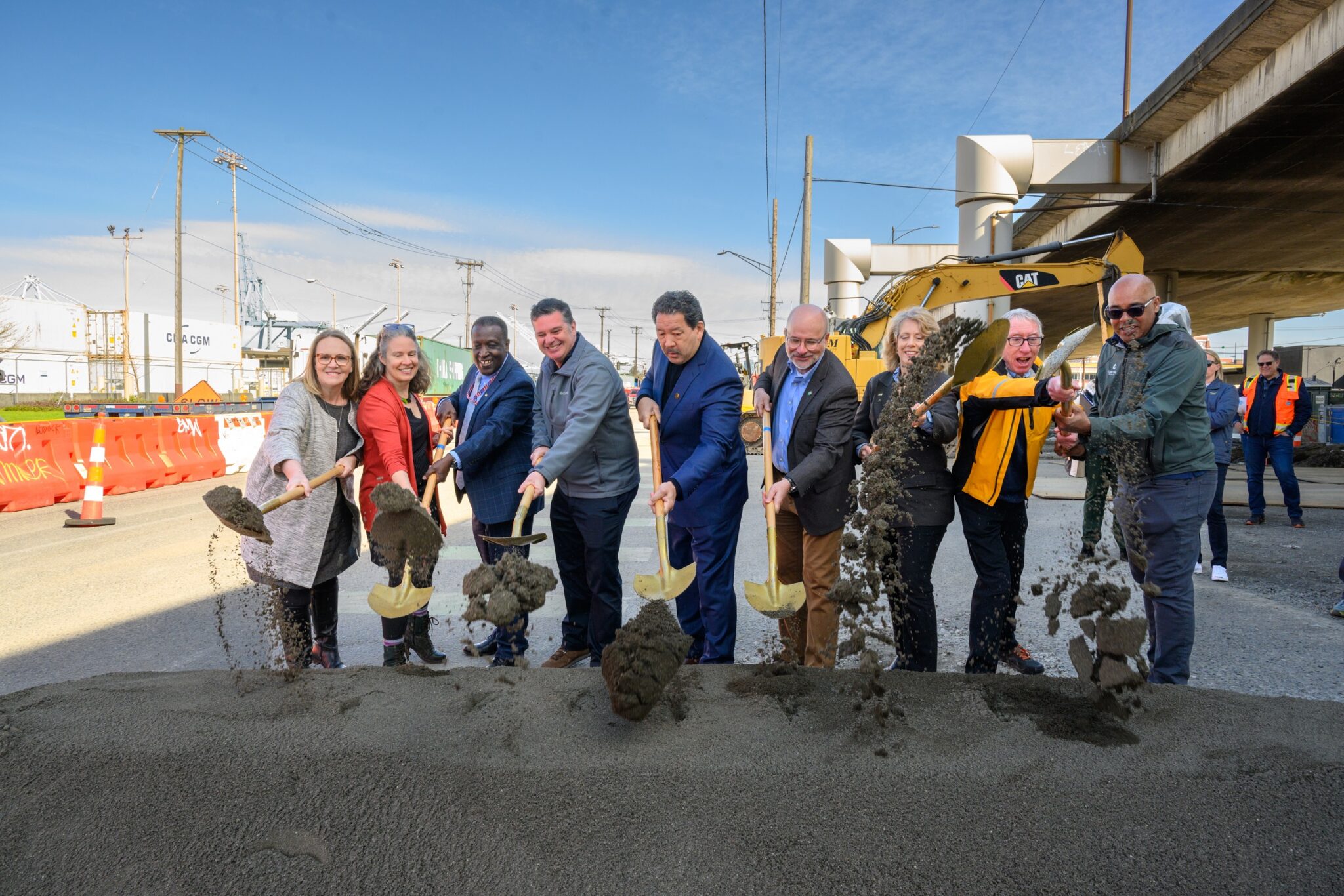 Several people use shovels at a groundbreaking event while smiling outdoors.