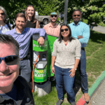 Several people smile on a sunny day standing next to an electric vehicle charging station.