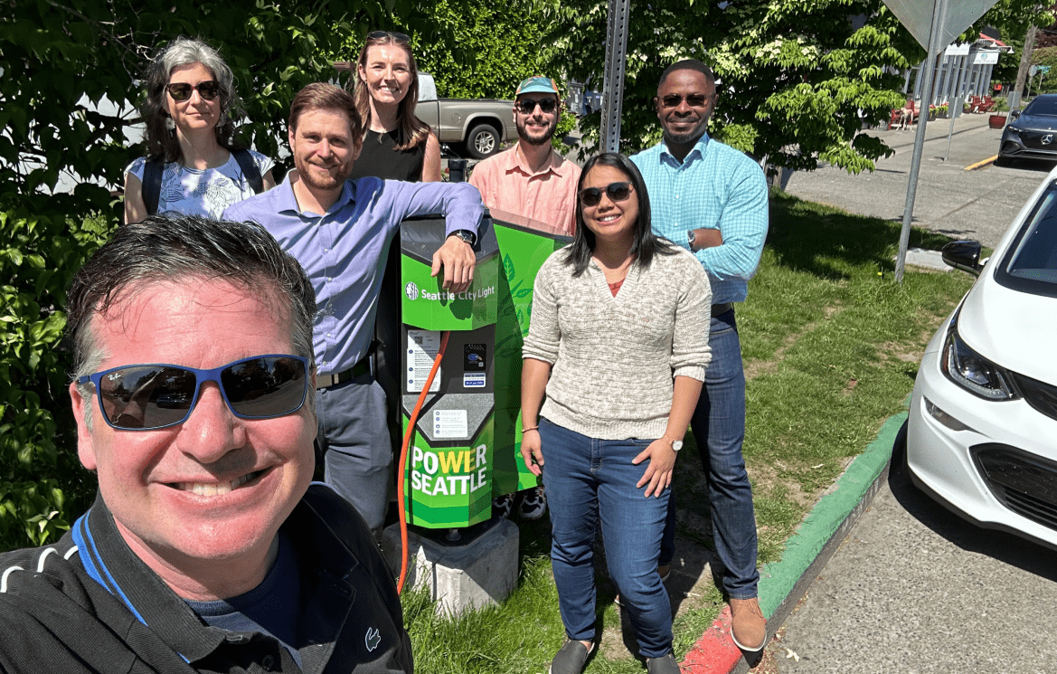 Several people smile on a sunny day standing next to an electric vehicle charging station.
