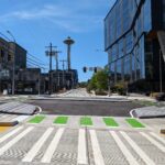 View of a crosswalk and bike path next to a street and large buildings with the Space Needle in the background.