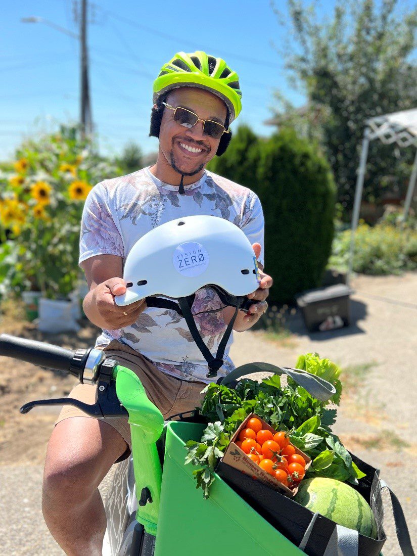 A person wearing a helmet sits on a bicycle while parked. Flowers are in the background and fresh produce in the bike's front basket.