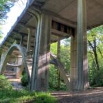 A large bridge viewed from below, looking up. Trees and vegetation are in the background.