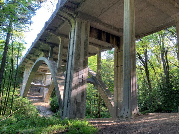 A large bridge viewed from below, looking up. Trees and vegetation are in the background.