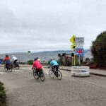 People bike along the street near a water body on a cloudy day.