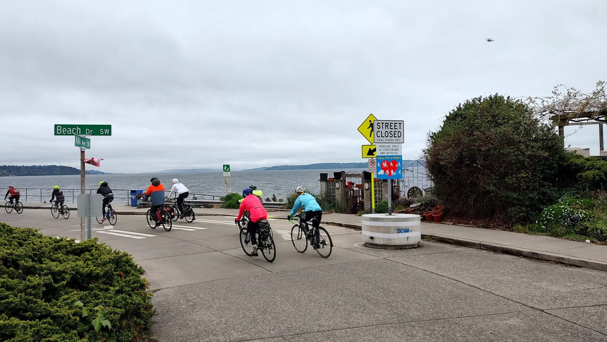 People bike along the street near a water body on a cloudy day.