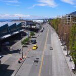 A photo of a street with large buildings to the left and trees to the right, on a sunny day. Several cars are traveling on the street.