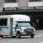 A shuttle bus leaves a building on a street. The words 'Seattle Children's Hospital' are on the building above the shuttle bus.