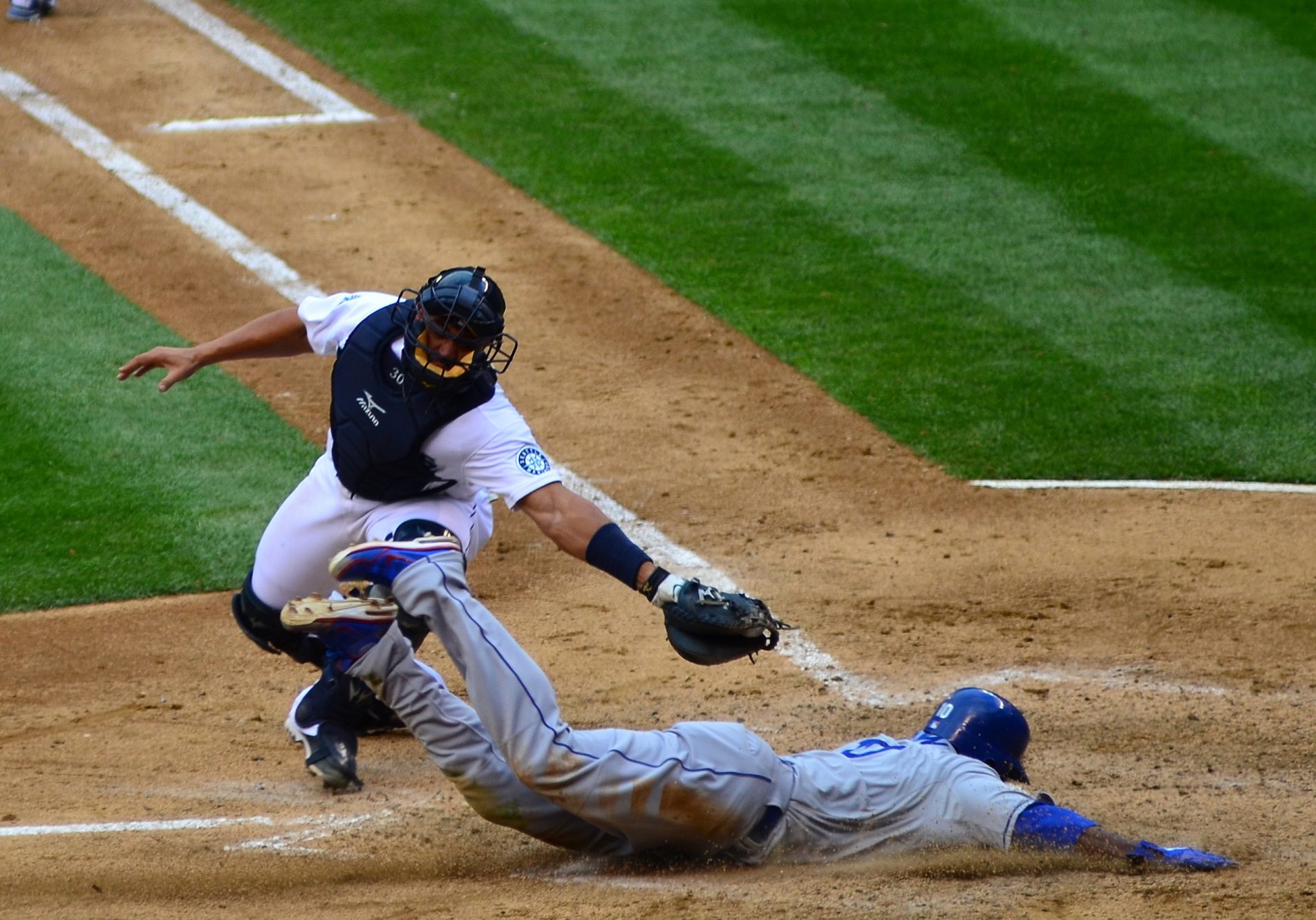 A catcher attempts to tag out a baserunner who is sliding into home plate during a baseball game. Green grass, brown dirt, and white lines are also in the image.