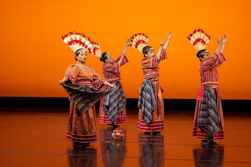 Four people dance on a stage as part of a performance. The background is a bright orange color.