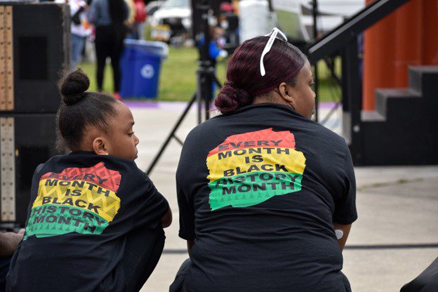 A Black girl and Black woman sit near a stage with a black shirt that writes "Every Month is Black History Month" on the back on top of a red, yellow, and green background.