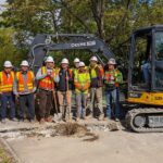 A large group of people stand in front of a heavy construction vehicle, wearing hard hats and safety vests on a sunny day. Trees are in the background.