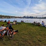 Several people sitting a large grassy hill overlooking a large body of water and a cityscape on a partly sunny day.