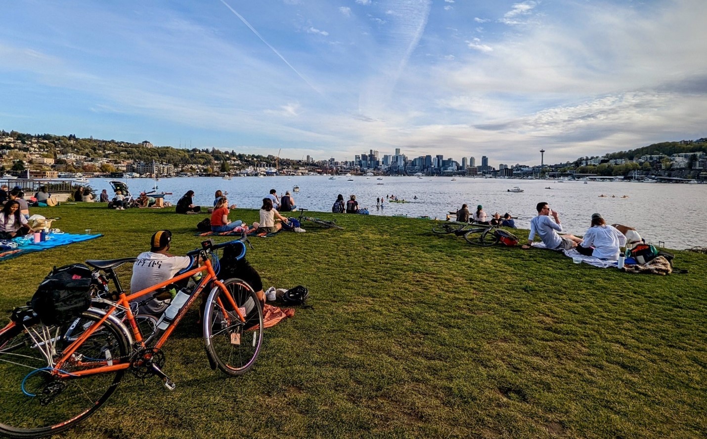 Several people sitting a large grassy hill overlooking a large body of water and a cityscape on a partly sunny day.