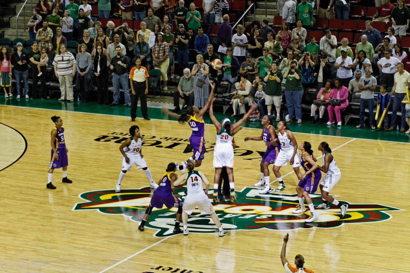 WNBA players tip off at the start of a basketball game. Many fans are in the background watching.
