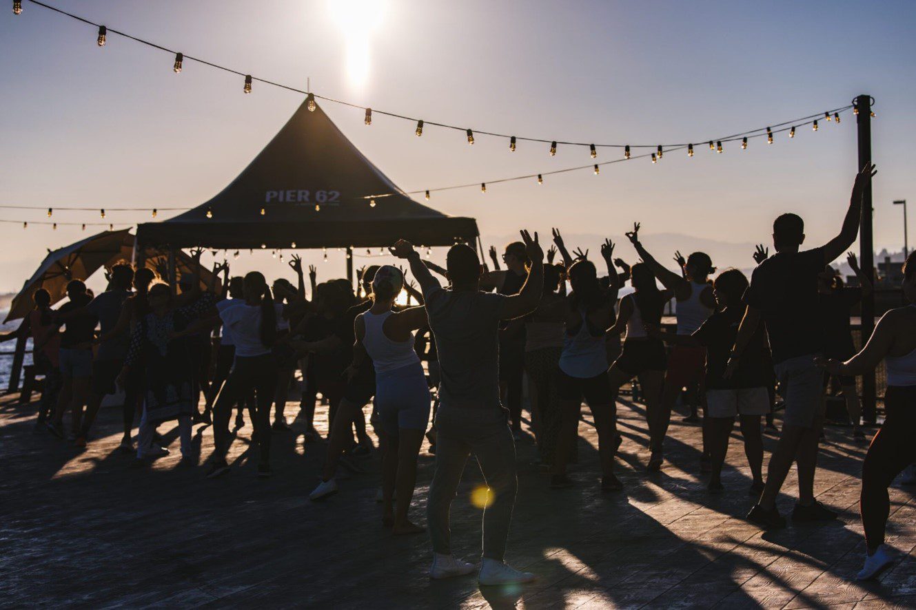 A large group of people celebrate and dance at a large celebration outdoors on a sunny day. A tent and strung lights are at the top and background.