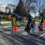 Several kids and adults cross the street with people wearing vests and holding orange flags helping them cross on a sunny day. A vehicle is in the background.