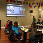 People gathered around a rectangular table shaped in an "L" as a video meeting display projects onto a screen at the front of the room. The room is decorated with colorful hexagons along the top towards the ceiling. A speaker presents on the left side behind a podium.