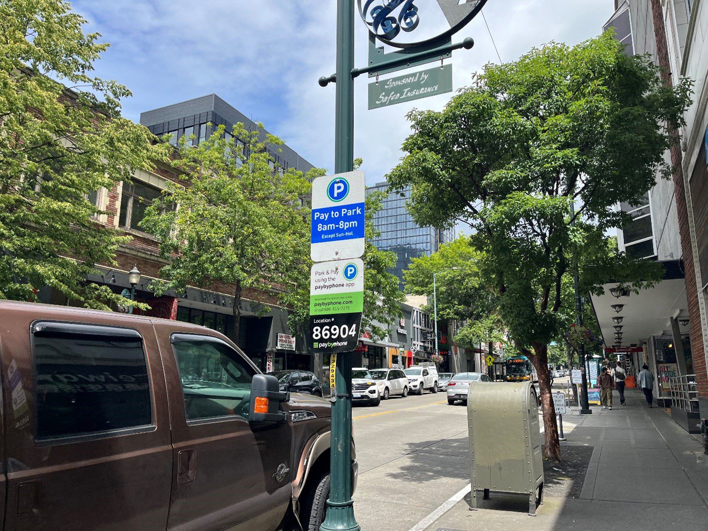 A truck parked on the street with large buildings and trees in the background. A parking sign is next to the truck.