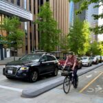 Man with black helmet and red flannel on black bike in the bike lane on the right hand side. Separated from a black car and a line of cars behind it with a road loaf. Brown and gray building in the background with trees planted on the edge of the sidewalks separating the road and sidewalk.