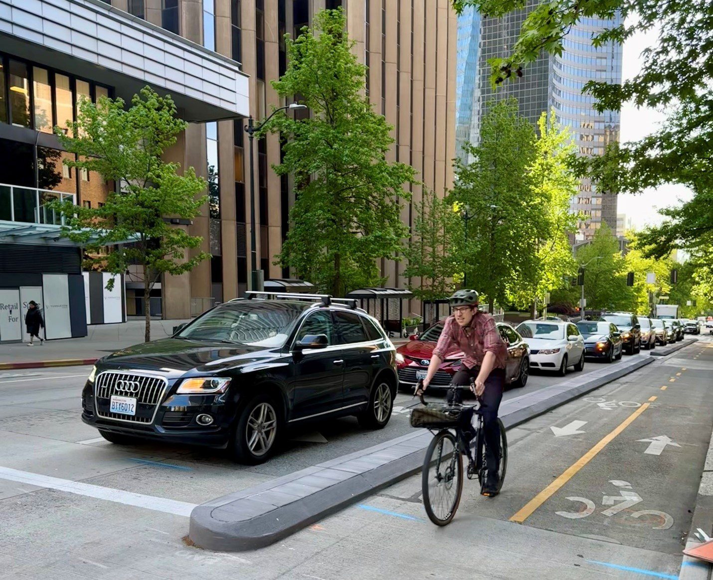 Man with black helmet and red flannel on black bike in the bike lane on the right hand side. Separated from a black car and a line of cars behind it with a road loaf. Brown and gray building in the background with trees planted on the edge of the sidewalks separating the road and sidewalk.