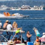 People in beach wear sitting next to a deep blue lake as a orange and white hydroplane zooms pass leaving a trail of water splashes behind it. Boats in the background also sit in the water.