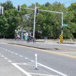 Two people bike in opposite directions. One female with a light blue helmet and pink and light grey tank and light grey leggings bikes to the left. One female in a white helmet, black top and leggings and a light blue back seat bikes to the right. Two red traffic lights above them and a bus stop in the middle of them in the background. The road takes up the bottom half of the image.