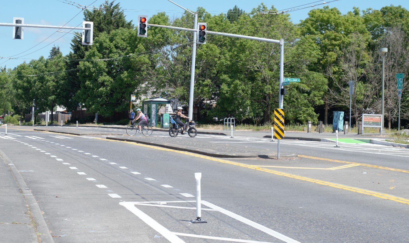 Two people bike in opposite directions. One female with a light blue helmet and pink and light grey tank and light grey leggings bikes to the left. One female in a white helmet, black top and leggings and a light blue back seat bikes to the right. Two red traffic lights above them and a bus stop in the middle of them in the background. The road takes up the bottom half of the image.