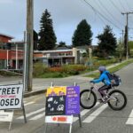 On a school morning. Gray and red-brown colored elementary school on the left and the road in front of it is marked with a street closed sign and school streets sign with yellow, black, and purple color blocks explaining school streets. A Kid bikes across the street at the crosswalk in a vibrant blue puffer jacket and light gray helmet. Students pull onto the sidewalk in front of the school with their bikes.