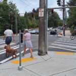 Man and women crossing to the left of an intersection. Women holding a black leash to a brown medium sized dog. A biker turns right at the intersection. Trees surround the intersection.