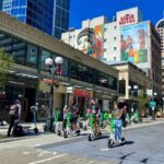 Sunny day photo of three friends riding bright neon green and white Lime scooters on the gray part of a pedestrian street. Beige two story building in the back and a white taller building peeking over behind it with colorful murals.