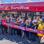 A large group of people gather around a ribbon in front of a bus that says "RapidRide" at the top as well as "G Downtown". the crowd holds large scissors as part of a ribbon cutting event.