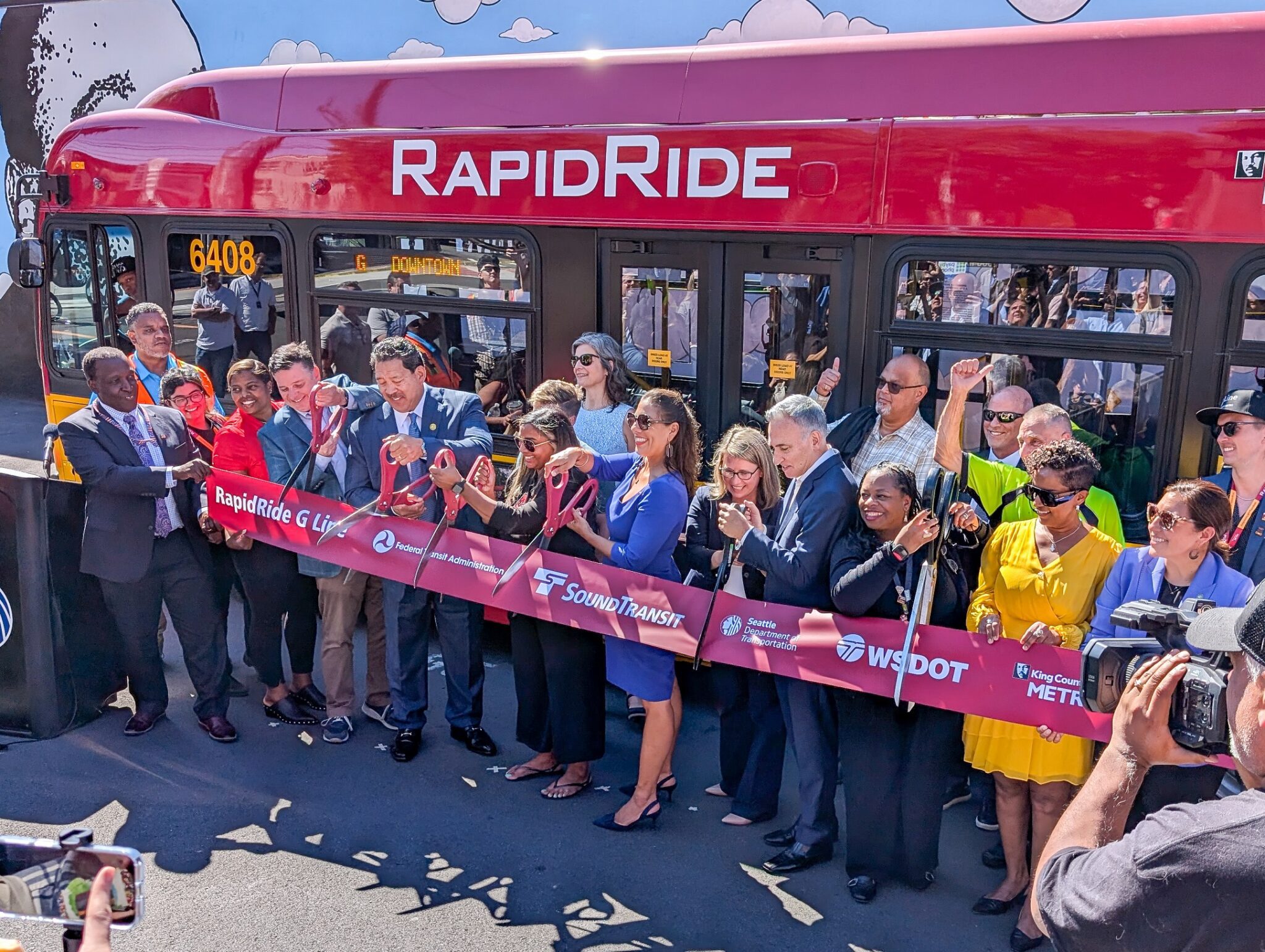 A large group of people gather around a ribbon in front of a bus that says 