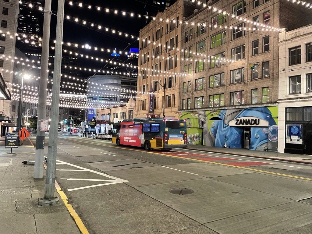 A bus travels along a street with strings of lights above at night. Large buildings and painted artwork on the walls are also in the image.