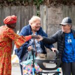 Three older adults smile while two of them shake hands. One woman is using a walker. They wear bright colorful clothing while standing outside at an event.