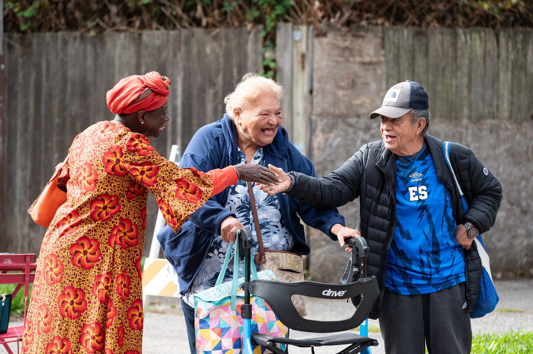 Three older adults smile while two of them shake hands. One woman is using a walker. They wear bright colorful clothing while standing outside at an event.