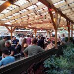 People sitting at an outdoor cafe seating under a light brown overhead structure that allows natural light in. People in image are playing chess on small chess boards. More than a dozen people are in the photo.