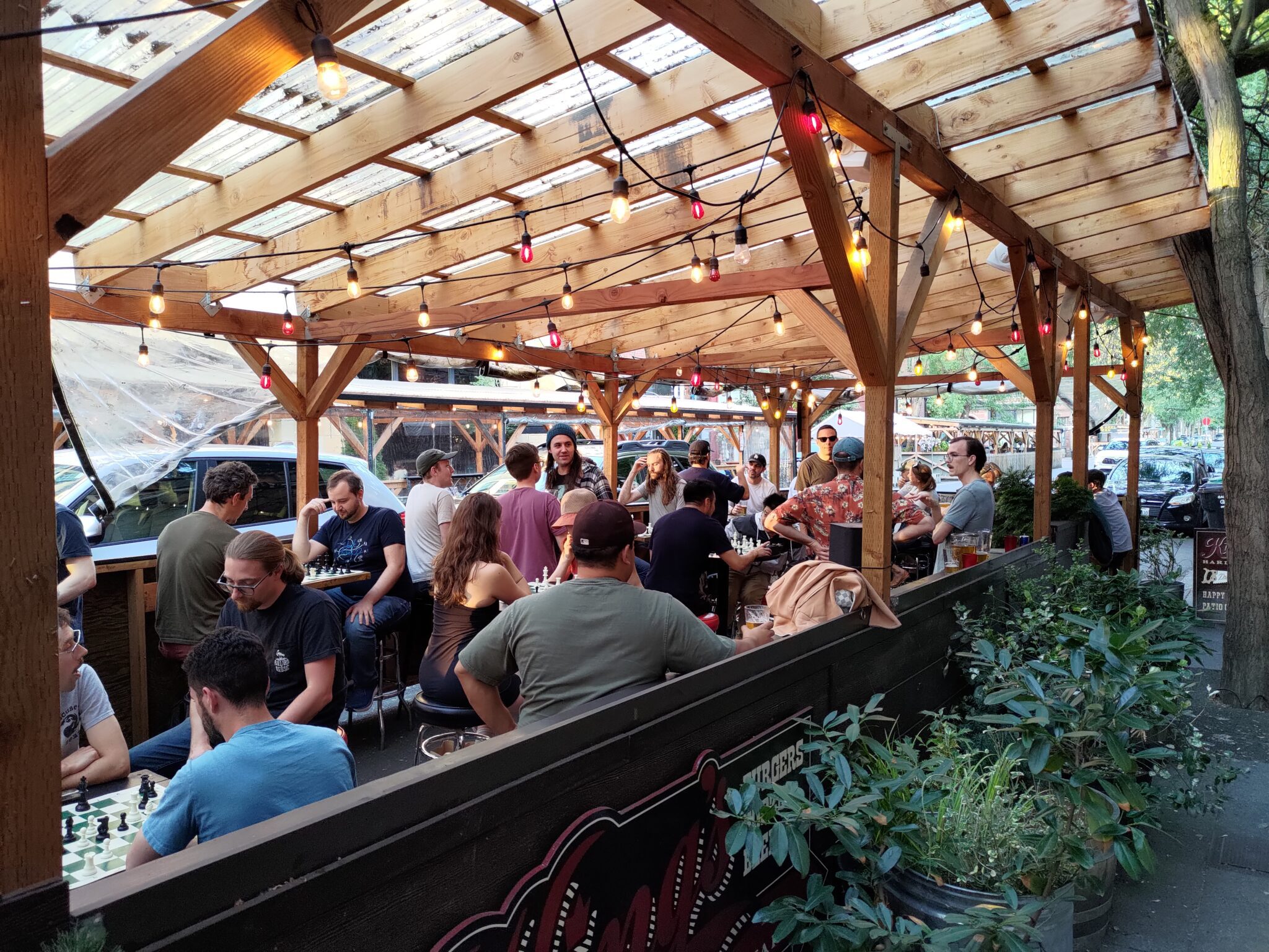 People sitting at an outdoor cafe seating under a light brown overhead structure that allows natural light in. People in image are playing chess on small chess boards. More than a dozen people are in the photo.