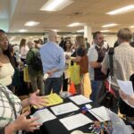 A group of people at a large indoor networking event. Three people connect while talking at the event. Handouts are on a table in the middle of the image.