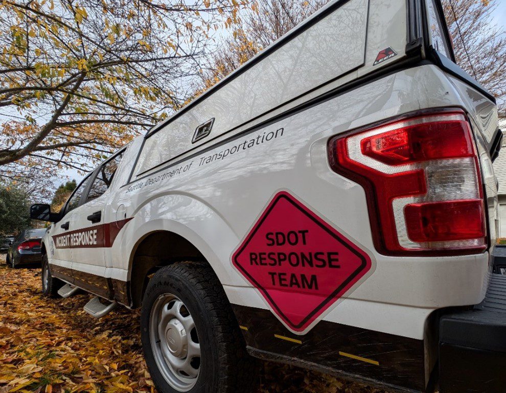 A white truck parked under trees and next to leaves. An orange logo says 