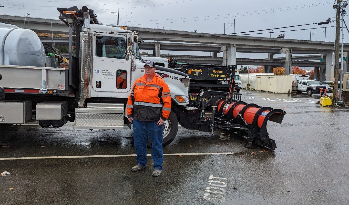 A person wearing an orange jacket stands in front of a large snow plow in an outdoor facility.