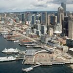 An aerial view of downtown Seattle with large buildings near the waterfront, and two ferry boats at a ferry terminal in the foreground.