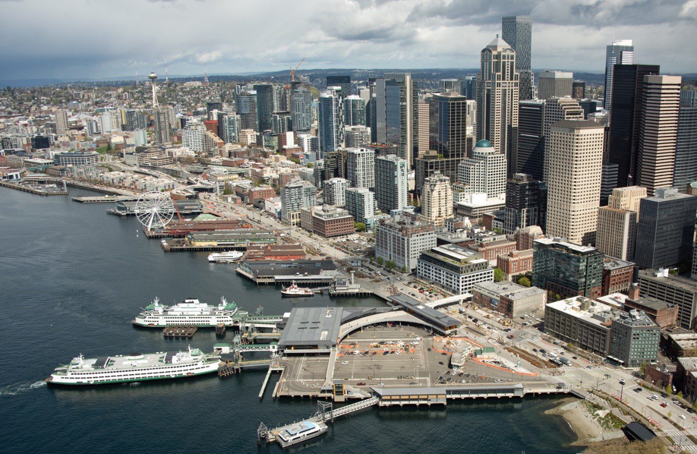 An aerial view of downtown Seattle with large buildings near the waterfront, and two ferry boats at a ferry terminal in the foreground.