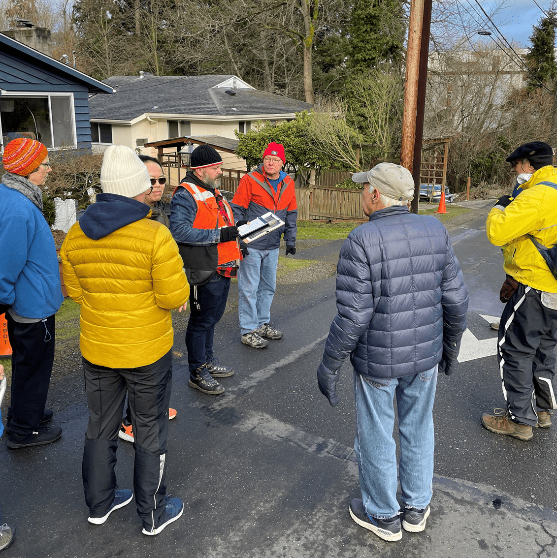 A group of several people stop while walking along a street in a residential neighborhood. One man is wearing an orange safety vest. Trees and houses are in the background.