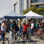 A large group of people celebrate biking at a community event outside. Two large canopy tents are in the background, with people next to bikes in the foreground.