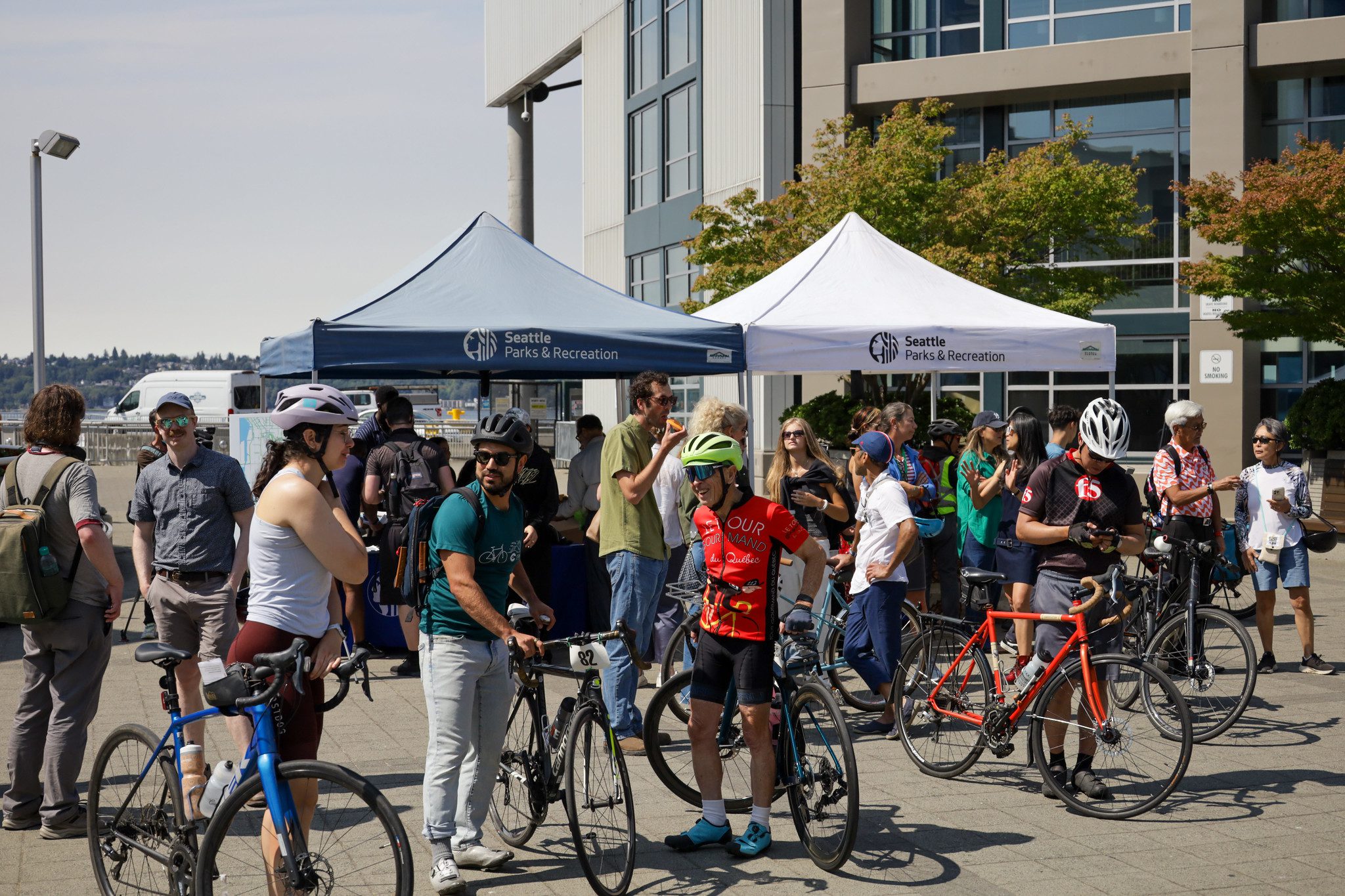 A large group of people celebrate biking at a community event outside. Two large canopy tents are in the background, with people next to bikes in the foreground.